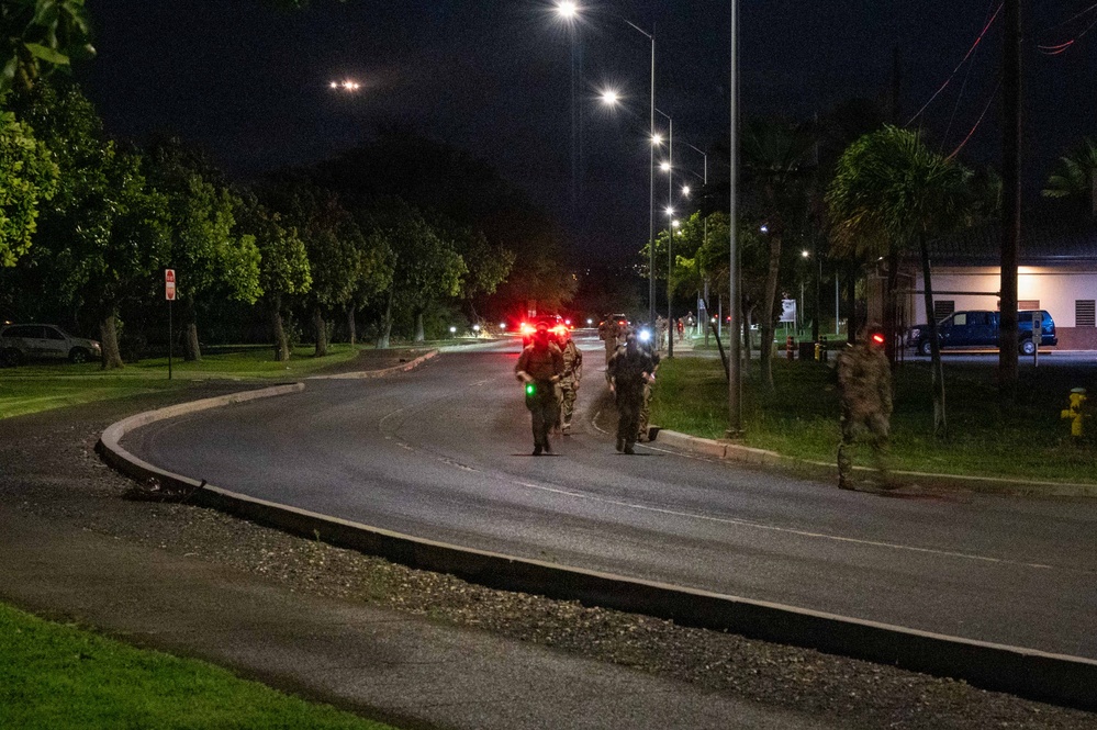 A nighttime scene showing a group of military or law enforcement personnel walking along a curved road, equipped with gear and visible light markers. The scene is illuminated by street lights and the personnel appear to be conducting a patrol or ruck march along what appears to be a military base or secured facility. Red and green light markers are visible on the personnel, consistent with tactical night movement indicators.
