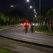 A nighttime scene showing a group of military or law enforcement personnel walking along a curved road, equipped with gear and visible light markers. The scene is illuminated by street lights and the personnel appear to be conducting a patrol or ruck march along what appears to be a military base or secured facility. Red and green light markers are visible on the personnel, consistent with tactical night movement indicators.