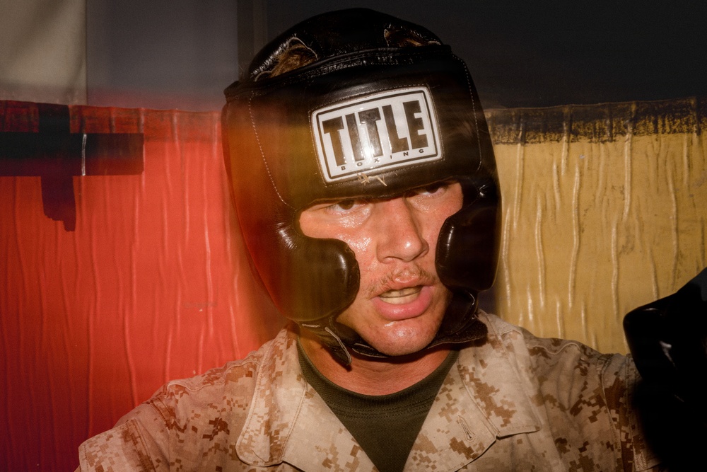 A man wearing a black 'Title Boxing' headguard and desert camouflage military uniform is photographed in close-up. His face shows signs of physical exertion, with visible sweat and a focused expression. The background features red and tan fabric or curtains, suggesting an informal or makeshift setting.