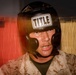 A man wearing a black 'Title Boxing' headguard and desert camouflage military uniform is photographed in close-up. His face shows signs of physical exertion, with visible sweat and a focused expression. The background features red and tan fabric or curtains, suggesting an informal or makeshift setting.