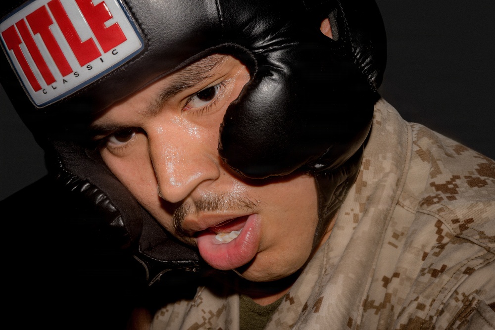A close-up photograph of a male individual wearing a black 'TITLE Classic' boxing headgear. He appears to be mid-activity, with his mouth open and tongue slightly visible, and his face shows signs of perspiration. He is wearing what appears to be a military camouflage uniform (MARPAT desert pattern), suggesting a military training or combatives context.