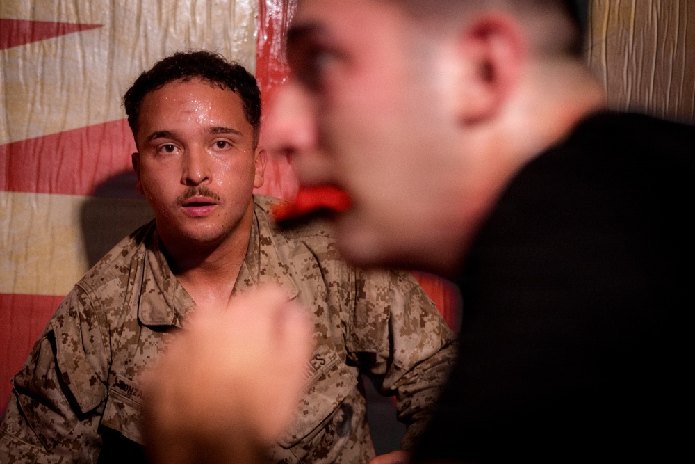 A military service member wearing desert camouflage (MARPAT) uniform is seated and appears to be in conversation with another individual in the foreground who is out of focus. The in-focus subject appears to be sweating and listening attentively. The background features what appears to be a fabric or tarp with red and white coloring.