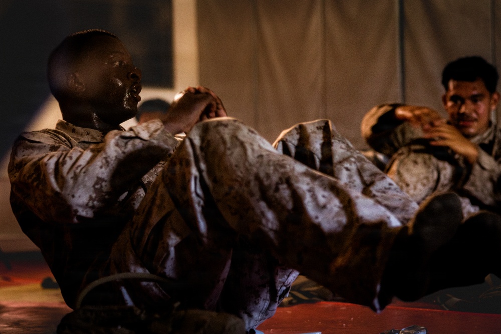 Two military personnel in camouflage uniforms are performing sit-up exercises, with their hands clasped behind their heads in standard physical training form. Their uniforms appear wet or soiled, suggesting an intense physical training exercise or field conditions. The setting appears to be a dimly lit area near a tent or temporary structure.