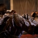 Two military personnel in camouflage uniforms are performing sit-up exercises, with their hands clasped behind their heads in standard physical training form. Their uniforms appear wet or soiled, suggesting an intense physical training exercise or field conditions. The setting appears to be a dimly lit area near a tent or temporary structure.