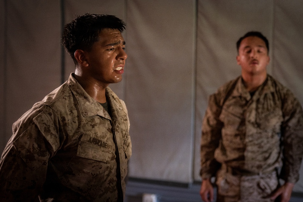 Two military personnel in desert camouflage uniforms are photographed in what appears to be a low-light indoor or covered outdoor setting near a tent or canvas structure. The foreground subject, whose name tape reads 'ANDRADE,' appears to be in physical distress or exertion, with a wet face and strained expression. The second individual stands in the background in a similar uniform, appearing calmer.
