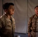 Two military personnel in desert camouflage uniforms are photographed in what appears to be a low-light indoor or covered outdoor setting near a tent or canvas structure. The foreground subject, whose name tape reads 'ANDRADE,' appears to be in physical distress or exertion, with a wet face and strained expression. The second individual stands in the background in a similar uniform, appearing calmer.