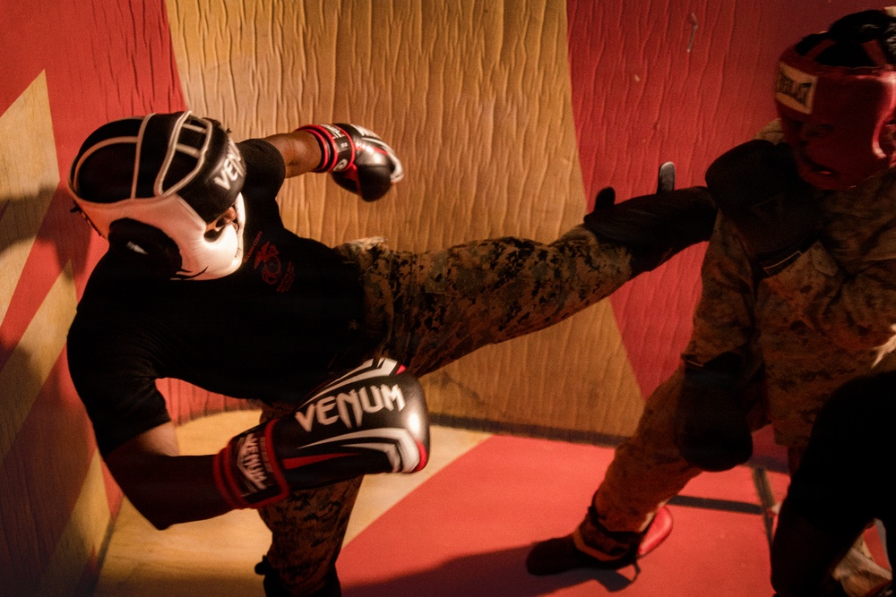 Two individuals wearing military camouflage (MARPAT pattern) uniforms are engaged in a sparring session in what appears to be a martial arts training facility. The left fighter is executing a high kick toward the opponent while wearing Venum-branded boxing gloves and headgear. Both participants are equipped with full protective gear including head guards and boxing gloves.
