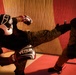 Two individuals wearing military camouflage (MARPAT pattern) uniforms are engaged in a sparring session in what appears to be a martial arts training facility. The left fighter is executing a high kick toward the opponent while wearing Venum-branded boxing gloves and headgear. Both participants are equipped with full protective gear including head guards and boxing gloves.