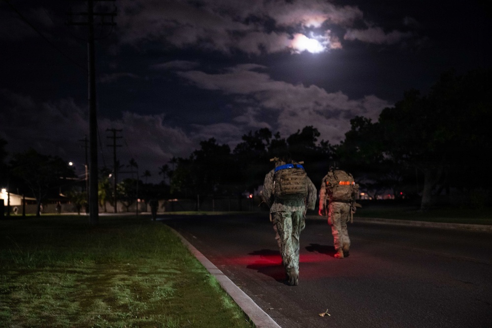 Two military personnel in full combat gear and tactical backpacks are conducting a nighttime ruck march along a paved road. The scene is illuminated by a bright moon partially obscured by clouds, along with red safety lights visible near the soldiers' feet. The setting appears to be a military installation or base, with utility poles, trees, and distant lighting visible in the background.