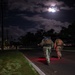 Two military personnel in full combat gear and tactical backpacks are conducting a nighttime ruck march along a paved road. The scene is illuminated by a bright moon partially obscured by clouds, along with red safety lights visible near the soldiers' feet. The setting appears to be a military installation or base, with utility poles, trees, and distant lighting visible in the background.