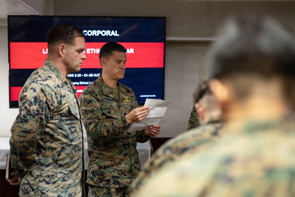 Military personnel in U.S. Marine Corps MARPAT camouflage uniforms are gathered in an indoor setting for a leadership training event. A central figure is reading from papers while standing in front of a presentation screen. The screen displays text indicating a 'Corporal Leadership and Ethics Seminar' course.