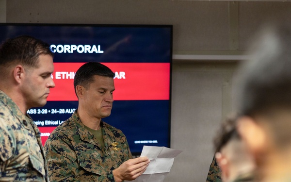 Military personnel in U.S. Marine Corps MARPAT camouflage uniforms are gathered in an indoor setting for a leadership training event. A central figure is reading from papers while standing in front of a presentation screen. The screen displays text indicating a 'Corporal Leadership and Ethics Seminar' course.