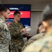 Military personnel in U.S. Marine Corps MARPAT camouflage uniforms are gathered in an indoor setting for a leadership training event. A central figure is reading from papers while standing in front of a presentation screen. The screen displays text indicating a 'Corporal Leadership and Ethics Seminar' course.