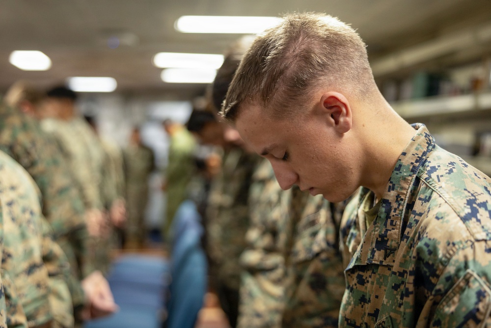 A group of military personnel in camouflage uniforms are gathered indoors with their heads bowed, suggesting a moment of prayer, reflection, or a moment of silence. The foreground features a young male service member in MARPAT (Marine pattern) digital camouflage with his head lowered. The setting appears to be an indoor facility, possibly a chapel, meeting room, or common area aboard a military installation or vessel.