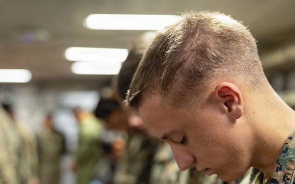 A group of military personnel in camouflage uniforms are gathered indoors with their heads bowed, suggesting a moment of prayer, reflection, or a moment of silence. The foreground features a young male service member in MARPAT (Marine pattern) digital camouflage with his head lowered. The setting appears to be an indoor facility, possibly a chapel, meeting room, or common area aboard a military installation or vessel.
