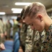 A group of military personnel in camouflage uniforms are gathered indoors with their heads bowed, suggesting a moment of prayer, reflection, or a moment of silence. The foreground features a young male service member in MARPAT (Marine pattern) digital camouflage with his head lowered. The setting appears to be an indoor facility, possibly a chapel, meeting room, or common area aboard a military installation or vessel.