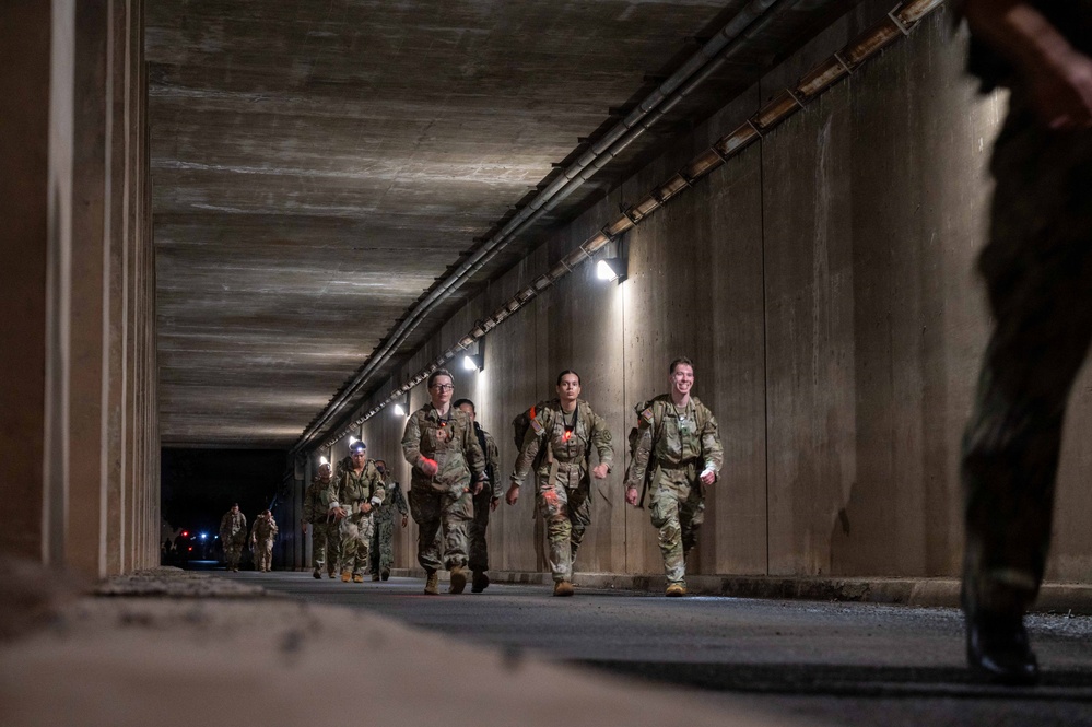 A group of military personnel in camouflage uniforms are marching through a concrete tunnel or underpass, carrying rucksacks and equipment. The scene appears to be a ruck march or training exercise conducted during nighttime or low-light conditions. Wall-mounted lights illuminate the tunnel, and several soldiers are wearing headlamps.