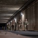 A group of military personnel in camouflage uniforms are marching through a concrete tunnel or underpass, carrying rucksacks and equipment. The scene appears to be a ruck march or training exercise conducted during nighttime or low-light conditions. Wall-mounted lights illuminate the tunnel, and several soldiers are wearing headlamps.