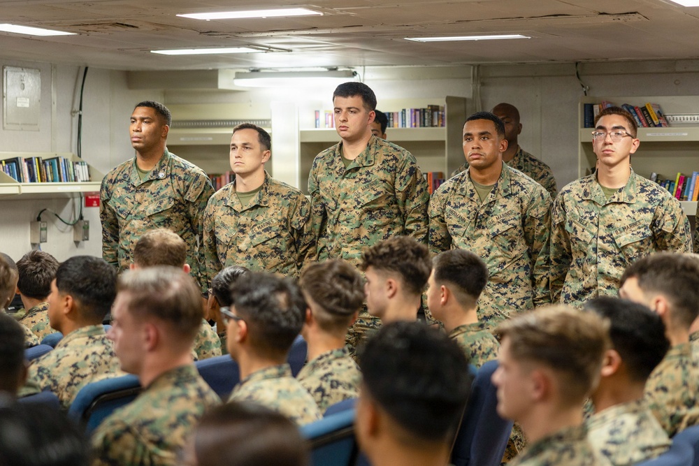 A group of U.S. Marines in MARPAT (Marine Pattern) camouflage uniforms are gathered in what appears to be a ship's library or briefing room. Five Marines are standing at the front facing an audience of seated Marines, suggesting a formal briefing, ceremony, or recognition event. The setting appears to be aboard a naval vessel based on the interior architecture.
