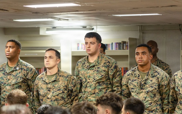 A group of U.S. Marines in MARPAT (Marine Pattern) camouflage uniforms are gathered in what appears to be a ship's library or briefing room. Five Marines are standing at the front facing an audience of seated Marines, suggesting a formal briefing, ceremony, or recognition event. The setting appears to be aboard a naval vessel based on the interior architecture.
