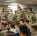 A group of U.S. Marines in MARPAT (Marine Pattern) camouflage uniforms are gathered in what appears to be a ship's library or briefing room. Five Marines are standing at the front facing an audience of seated Marines, suggesting a formal briefing, ceremony, or recognition event. The setting appears to be aboard a naval vessel based on the interior architecture.