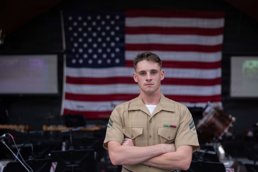 A young male service member in a tan/khaki military uniform stands with arms crossed in front of a large American flag. He is wearing service ribbons on his left chest and rank insignia on his collar. The setting appears to be an indoor venue or stage with musical instruments visible in the background.