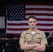 A young male service member in a tan/khaki military uniform stands with arms crossed in front of a large American flag. He is wearing service ribbons on his left chest and rank insignia on his collar. The setting appears to be an indoor venue or stage with musical instruments visible in the background.