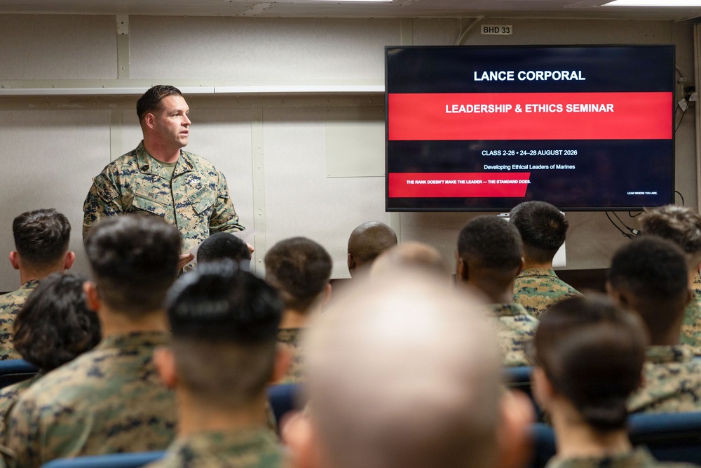 A military instructor in U.S. Marine Corps camouflage uniform is presenting at a Lance Corporal Leadership &amp; Ethics Seminar to a room full of seated Marines. The presentation slide identifies this as Class 2-26, scheduled for 24-28 August 2026, focused on developing ethical leaders. The setting appears to be a formal military classroom or briefing room.
