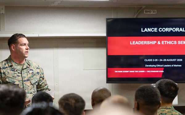 A military instructor in U.S. Marine Corps camouflage uniform is presenting at a Lance Corporal Leadership &amp; Ethics Seminar to a room full of seated Marines. The presentation slide identifies this as Class 2-26, scheduled for 24-28 August 2026, focused on developing ethical leaders. The setting appears to be a formal military classroom or briefing room.