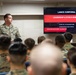 A military instructor in U.S. Marine Corps camouflage uniform is presenting at a Lance Corporal Leadership &amp; Ethics Seminar to a room full of seated Marines. The presentation slide identifies this as Class 2-26, scheduled for 24-28 August 2026, focused on developing ethical leaders. The setting appears to be a formal military classroom or briefing room.