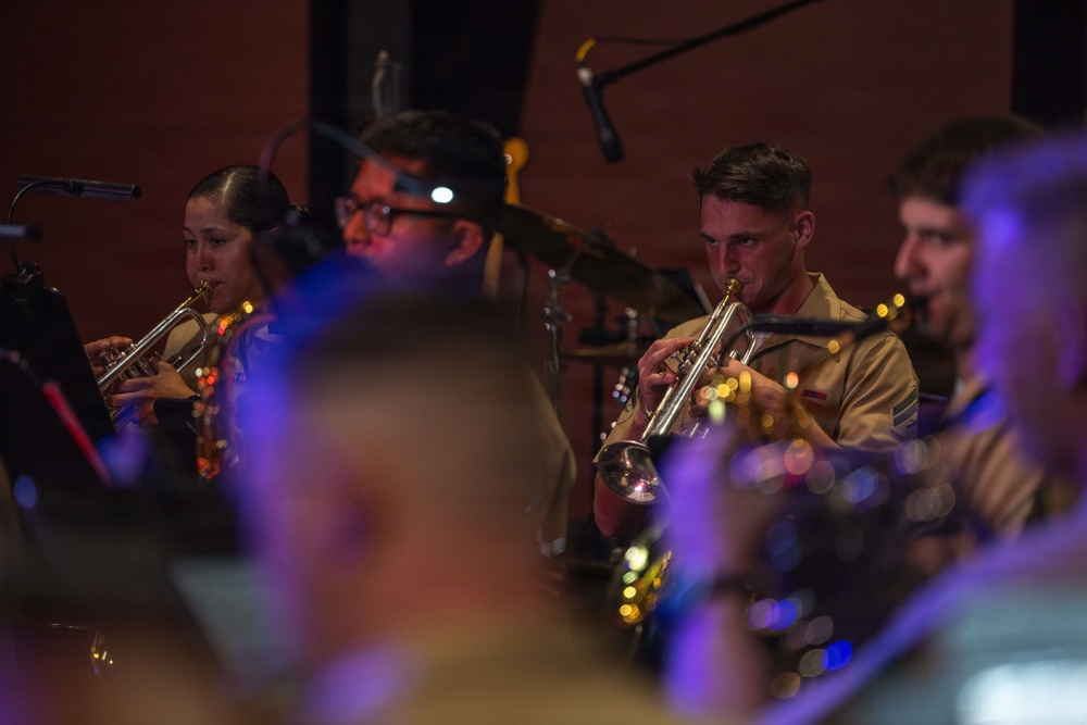A military band performance is captured with multiple musicians playing brass instruments, specifically trumpets. The performers are wearing tan/khaki military uniforms and are seated in an ensemble setting with music stands and stage lighting. The image has a shallow depth of field with blurred foreground elements framing the in-focus musicians.