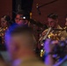 A military band performance is captured with multiple musicians playing brass instruments, specifically trumpets. The performers are wearing tan/khaki military uniforms and are seated in an ensemble setting with music stands and stage lighting. The image has a shallow depth of field with blurred foreground elements framing the in-focus musicians.