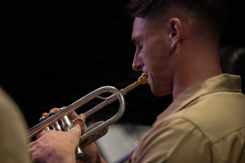 A close-up profile shot of a young man in a tan/khaki military-style uniform playing a silver flugelhorn or trumpet with a gold-colored mouthpiece. The image is tightly framed, emphasizing the musician's focused expression and the instrument's metallic details. The dark background creates strong contrast, highlighting the subject.