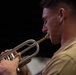 A close-up profile shot of a young man in a tan/khaki military-style uniform playing a silver flugelhorn or trumpet with a gold-colored mouthpiece. The image is tightly framed, emphasizing the musician's focused expression and the instrument's metallic details. The dark background creates strong contrast, highlighting the subject.