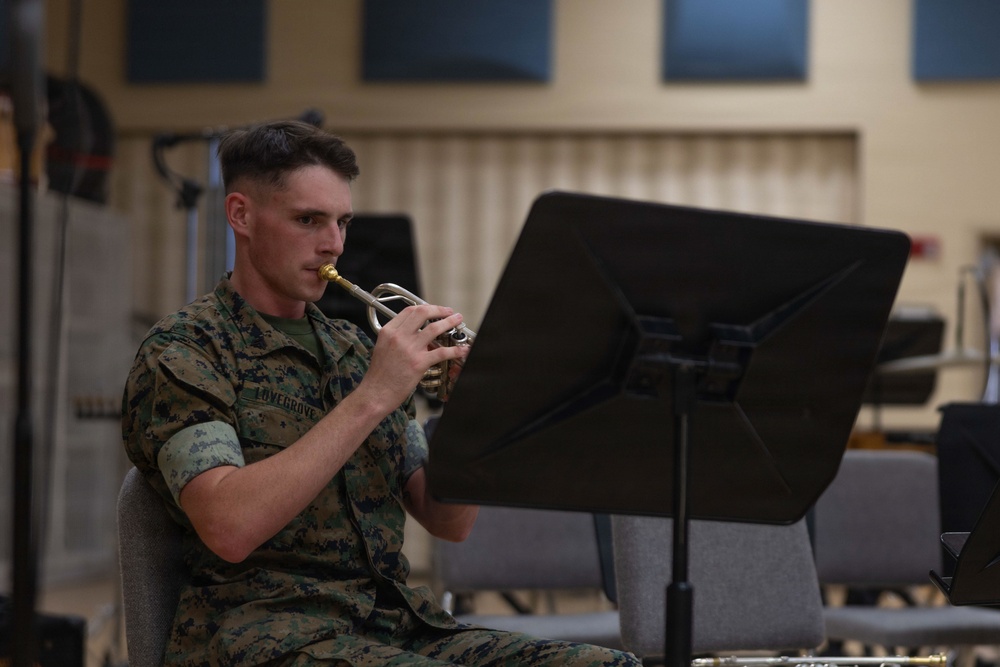 A U.S. Marine in digital camouflage uniform is seated and playing a trumpet or bugle while reading sheet music from a black music stand. The setting appears to be an indoor rehearsal or band room, identifiable by acoustic foam panels on the walls and rows of empty chairs in the background. The name tape on the uniform reads 'LOVEGROVE'.