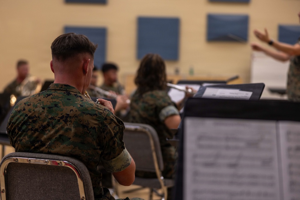 A group of military personnel in camouflage uniforms are participating in a band rehearsal in what appears to be an acoustically treated rehearsal room. Musicians are seated with music stands holding sheet music, while a conductor is visible in the background directing the ensemble. The foreground shows a musician holding a brass instrument with sheet music visible on a stand.