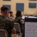A group of military personnel in camouflage uniforms are participating in a band rehearsal in what appears to be an acoustically treated rehearsal room. Musicians are seated with music stands holding sheet music, while a conductor is visible in the background directing the ensemble. The foreground shows a musician holding a brass instrument with sheet music visible on a stand.