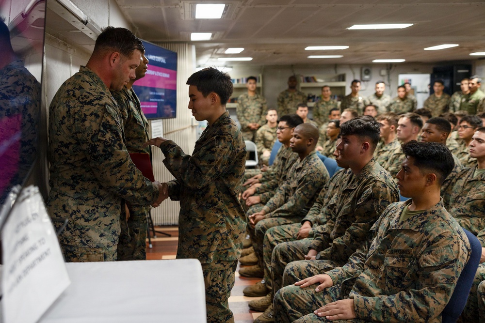 A military ceremony is taking place in an indoor briefing or assembly room, where a senior Marine officer is shaking hands with and apparently presenting something to a junior service member. A large audience of uniformed Marines in MARPAT digital camouflage uniforms are seated and observing the proceedings. The setting appears to be aboard a naval vessel or military installation.