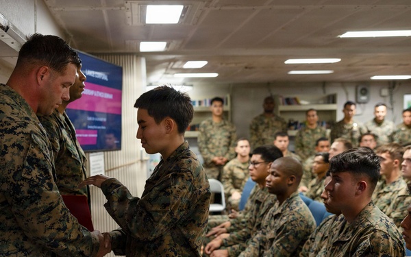 A military ceremony is taking place in an indoor briefing or assembly room, where a senior Marine officer is shaking hands with and apparently presenting something to a junior service member. A large audience of uniformed Marines in MARPAT digital camouflage uniforms are seated and observing the proceedings. The setting appears to be aboard a naval vessel or military installation.