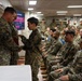 A military ceremony is taking place in an indoor briefing or assembly room, where a senior Marine officer is shaking hands with and apparently presenting something to a junior service member. A large audience of uniformed Marines in MARPAT digital camouflage uniforms are seated and observing the proceedings. The setting appears to be aboard a naval vessel or military installation.