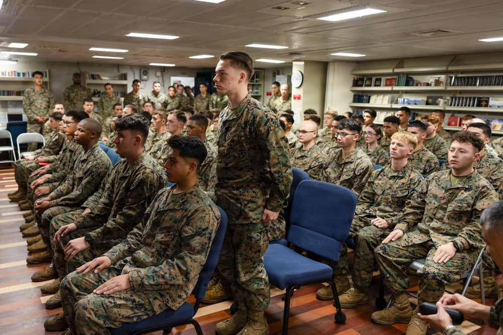 A large group of U.S. Marines in MARPAT camouflage uniforms are assembled in what appears to be a ship's library or briefing room. Most personnel are seated in rows of blue chairs, while one Marine stands at the center, possibly responding to a question or being recognized. The setting suggests a formal briefing, all-hands meeting, or command presentation aboard a naval vessel.