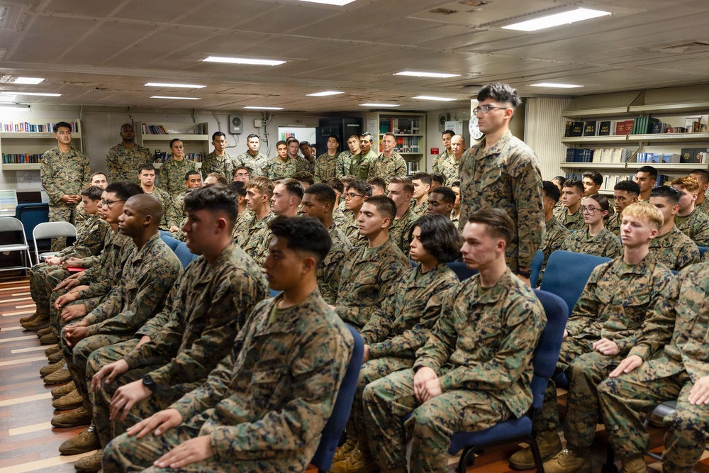 A large group of military personnel in woodland camouflage uniforms (MARPAT pattern consistent with U.S. Marines) are assembled in what appears to be a shipboard library or briefing room. The personnel are seated in rows of blue chairs and standing along the walls, appearing to attend a briefing or formation. One individual is standing among the seated group, possibly addressing or responding to a speaker.
