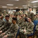 A large group of military personnel in woodland camouflage uniforms (MARPAT pattern consistent with U.S. Marines) are assembled in what appears to be a shipboard library or briefing room. The personnel are seated in rows of blue chairs and standing along the walls, appearing to attend a briefing or formation. One individual is standing among the seated group, possibly addressing or responding to a speaker.