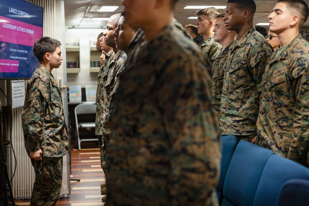 A group of U.S. Marines in MARPAT camouflage uniforms are attending a Lance Corporal Leadership &amp; Ethics Seminar, standing at attention in what appears to be a library or classroom setting. A television screen displays the seminar title slide with details including 'Class 3-20' and dates in August 2020, with the subtitle 'Developing Ethical Leaders of Marines.' One Marine on the left appears to be addressing or presenting to the group.