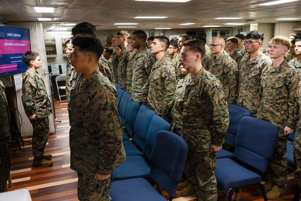 A group of U.S. Marines in woodland MARPAT camouflage uniforms are standing at attention in a seminar room. A presentation screen on the left displays 'Lance Corporal Leadership &amp; Ethics Seminar.' The setting appears to be an indoor military training or briefing facility with blue chairs arranged in rows.