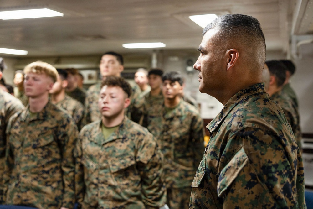 A group of military personnel wearing MARPAT (Marine Pattern) digital camouflage uniforms are assembled in what appears to be an interior space of a naval vessel or military facility. A senior Marine in the foreground is facing the assembled group, suggesting a briefing, inspection, or formation is taking place. The scene depicts a formal military gathering with multiple service members standing at attention or in a relaxed formation.