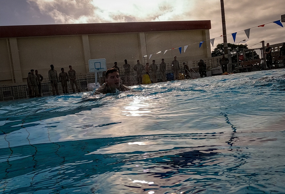 A person in what appears to be military uniform or clothing is swimming in an outdoor pool, likely during a military water survival training exercise. A large group of uniformed personnel are standing poolside observing the activity. The setting includes a basketball court backdrop, decorative bunting flags, and an overcast sky.