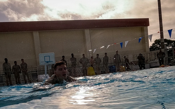 A person in what appears to be military uniform or clothing is swimming in an outdoor pool, likely during a military water survival training exercise. A large group of uniformed personnel are standing poolside observing the activity. The setting includes a basketball court backdrop, decorative bunting flags, and an overcast sky.