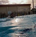 A person in what appears to be military uniform or clothing is swimming in an outdoor pool, likely during a military water survival training exercise. A large group of uniformed personnel are standing poolside observing the activity. The setting includes a basketball court backdrop, decorative bunting flags, and an overcast sky.