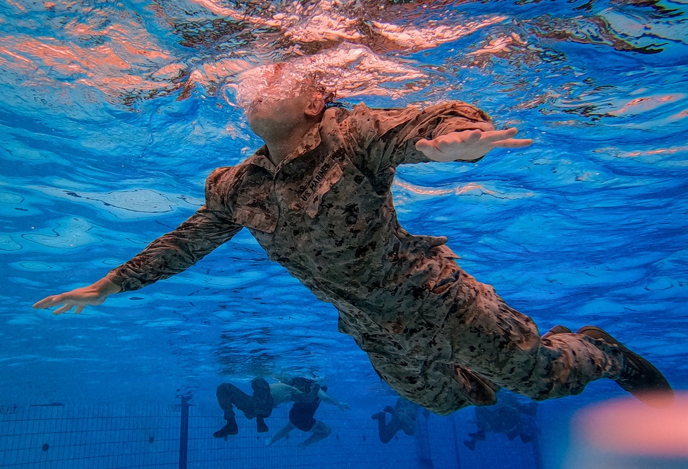An underwater photograph taken inside a swimming pool shows a U.S. Marine in full camouflage uniform (MARPAT pattern) swimming beneath the surface. The image is captured from below, providing a unique perspective looking up toward the water surface. Additional personnel in uniform are visible in the background at the pool bottom, suggesting a military water survival or combat swimmer training exercise.